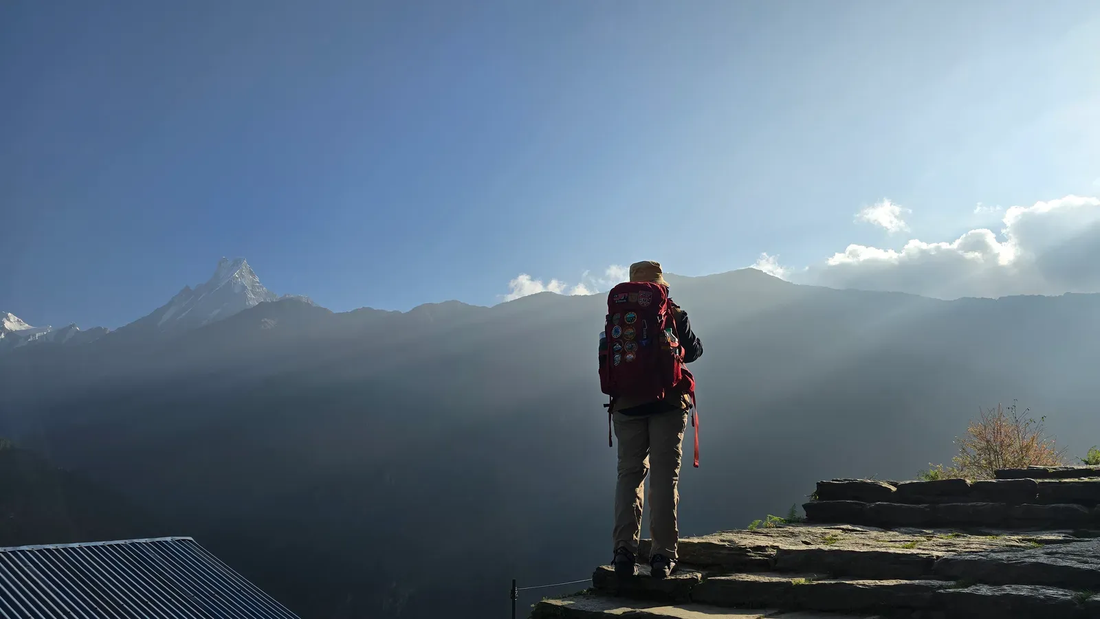 A trekker at a teahouse viewpoint facing Machapuchhre (Fishtail) at dawn, sun breaking over the ridge and clear sky above the peak