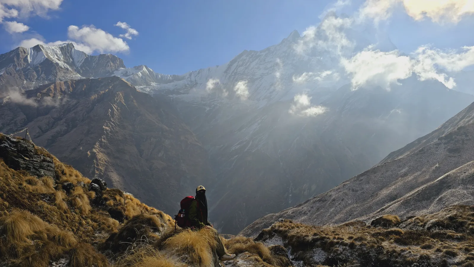 A trekker resting on a high grassy ridge, snow peaks and swirling cloud above on the approach to Machhapuchhre Base Camp
