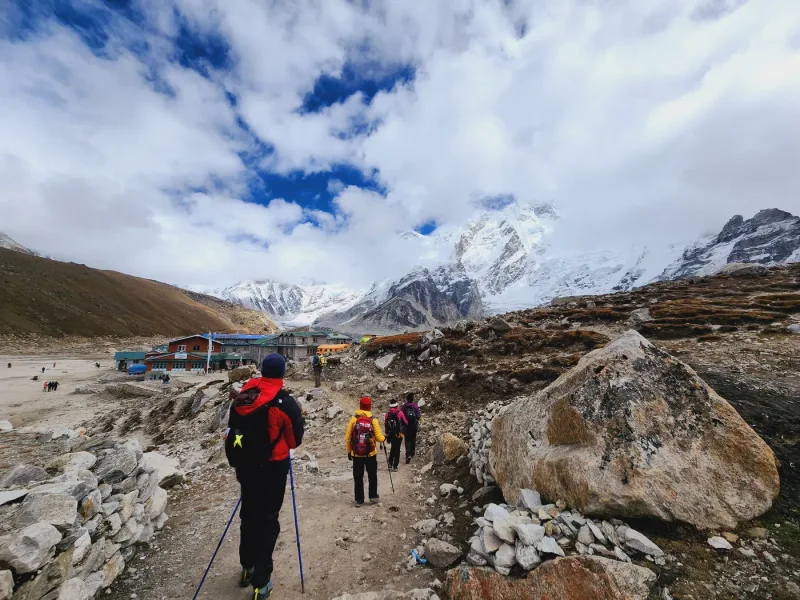 A group of trekkers strung along the trail toward a high teahouse settlement, snow peaks behind